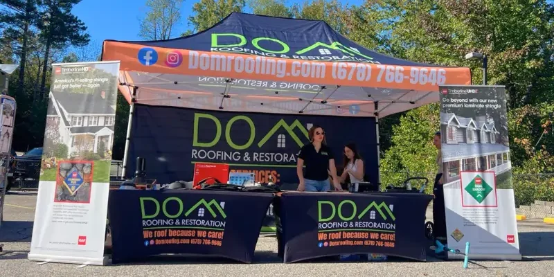 Roof Replacement 6 The Dom Roofing & Restoration event booth, featuring a branded tent and banners, set up to showcase their roofing services to the local community.